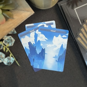 May include: Three blue and white "Island" cards with a landscape illustration of mountains, clouds, and water. The cards are stacked on a dark surface, with a few other items in the background.