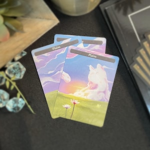 May include: Three "Plains" cards with a landscape illustration of a sunset over a field of grass and flowers. The sky is filled with clouds in shades of blue, pink, and purple. The cards are stacked on a dark surface.