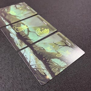 Op de afbeelding: Drie groene en blauwe moeraskaarten met een gloeiend katten silhouet op elke kaart. De kaarten hebben het woord "Moeras" erop gedrukt.