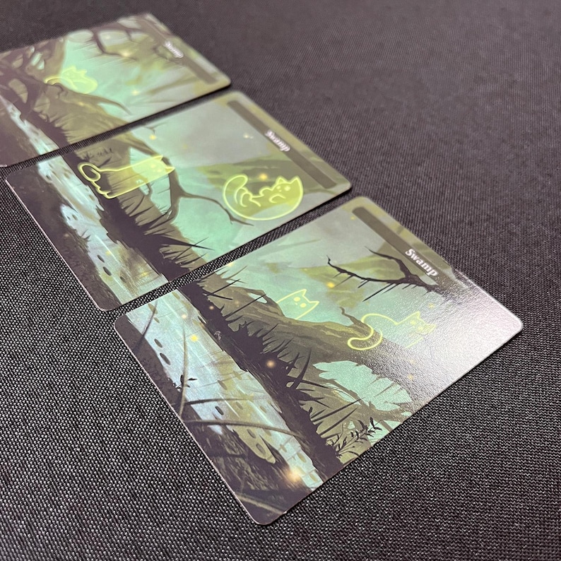 Op de afbeelding: Drie groene en blauwe moeraskaarten met een gloeiend katten silhouet op elke kaart. De kaarten hebben het woord "Moeras" erop gedrukt.