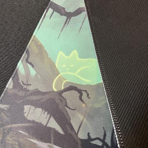 May include: A close-up of a dark green and black fabric with a glowing green cat silhouette on a branch. The fabric has a subtle texture and a white seam along the edge.