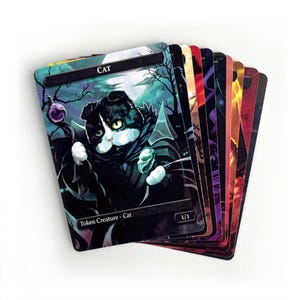 May include: A stack of fantasy trading cards featuring a black and white cat character with a cape and a staff. The card is labeled "CAT" and "Token Creature - Cat". Other cards in the stack have colorful fantasy art.