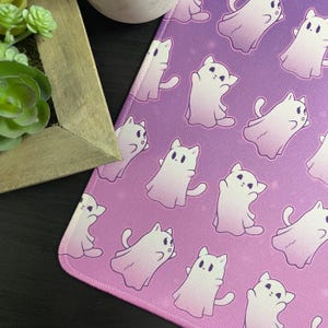 Peut inclure: Un tapis de souris à dégradé rose et violet avec un motif répétitif de chats fantômes blancs. Les chats sont dans une variété de poses, y compris assis, debout et flottant. Le tapis de souris a un bord cousu.
