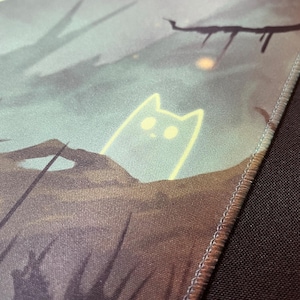 May include: A close-up of a green and brown fabric mousepad with a stitched edge. The mousepad features a light yellow outline of a cat with large eyes.