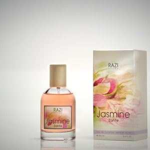 May include: A glass bottle of Jasmine Zante perfume by Razi Aroma. The bottle has a wooden cap and is filled with a light pink liquid. The label is white with pink and green floral designs and the text "RAZI Aroma Jasmine Zante Eau de Toilette Natural Spray 100 ml 3.4 fl oz."
