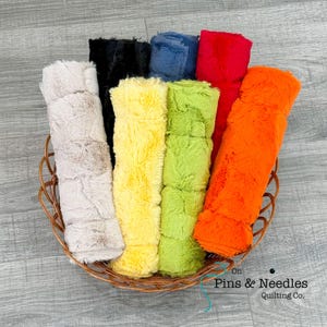 May include: A collection of seven plush, faux fur fabric rolls in beige, black, blue, red, yellow, lime green, and orange, arranged upright in a brown woven basket. The text "On Pins & Needles Quilting Co." is printed in the bottom right corner.
