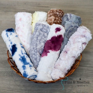 May include: A wicker basket holds a collection of plush, rolled fabric pieces in various colors. The fabrics include white with blue tie-dye, white with pink spots, yellow, brown, gray, and white with burgundy spots. The fabrics are soft and fluffy.