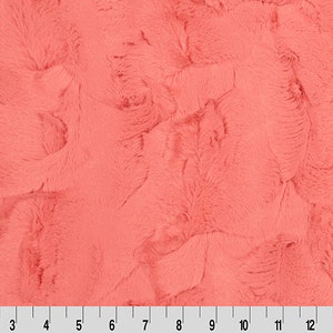May include: A close-up of a coral-coloured faux fur fabric. The fabric has a soft, plush texture and a subtle swirl pattern.