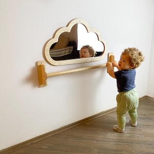 Könnte beinhalten: Ein hölzerner, wolkenförmiger Spiegel mit dem Spiegelbild eines Kindes. Unter dem Spiegel ist eine Holzstange an der Wand befestigt. Ein Kind steht vor dem Spiegel und greift nach der Stange.