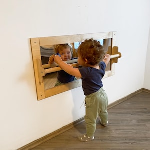 Puede incluir: Un niño se para frente a un espejo con marco de madera y una barra de madera unida. El espejo está montado en una pared blanca. El niño está mirando su reflejo en el espejo.
