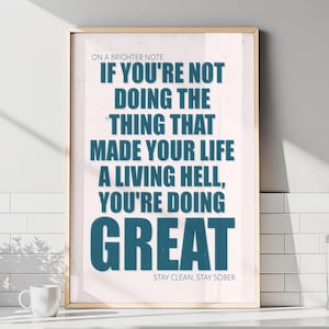 May include: A framed poster with a gold-colored frame. The poster has a light pink background with teal text that reads, "IF YOU'RE NOT DOING THE THING THAT MADE YOUR LIFE A LIVING HELL, YOU'RE DOING GREAT." The bottom text reads, "STAY CLEAN. STAY SOBER."