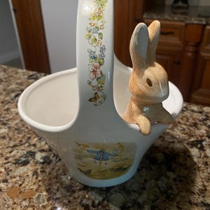 May include: A white ceramic basket featuring a sculpted light brown rabbit perched on the rim. The handle is decorated with a floral and butterfly pattern, while the front of the basket displays a colorful illustration of a scarecrow.