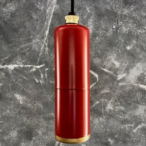 Op de afbeelding: Een rode hanglamp in de vorm van een cilinder met een gouden boven- en onderkant. De lamp hangt aan een zwart snoer tegen een grijze, getextureerde achtergrond.