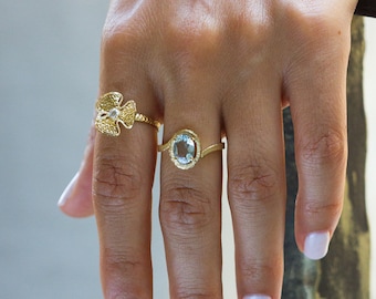 May include: Two gold rings on a hand. The ring on the index finger has a light blue oval gemstone. The ring on the middle finger has a floral design with a small white gemstone in the centre.