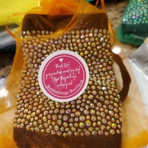 May include: A brown pouch with a sparkly design is in a bright orange organza bag. The pouch has a round sticker with the text "Thank You! Your purchase made my day! I hope the package makes you! Baddablingz Boutique" in pink and white text.