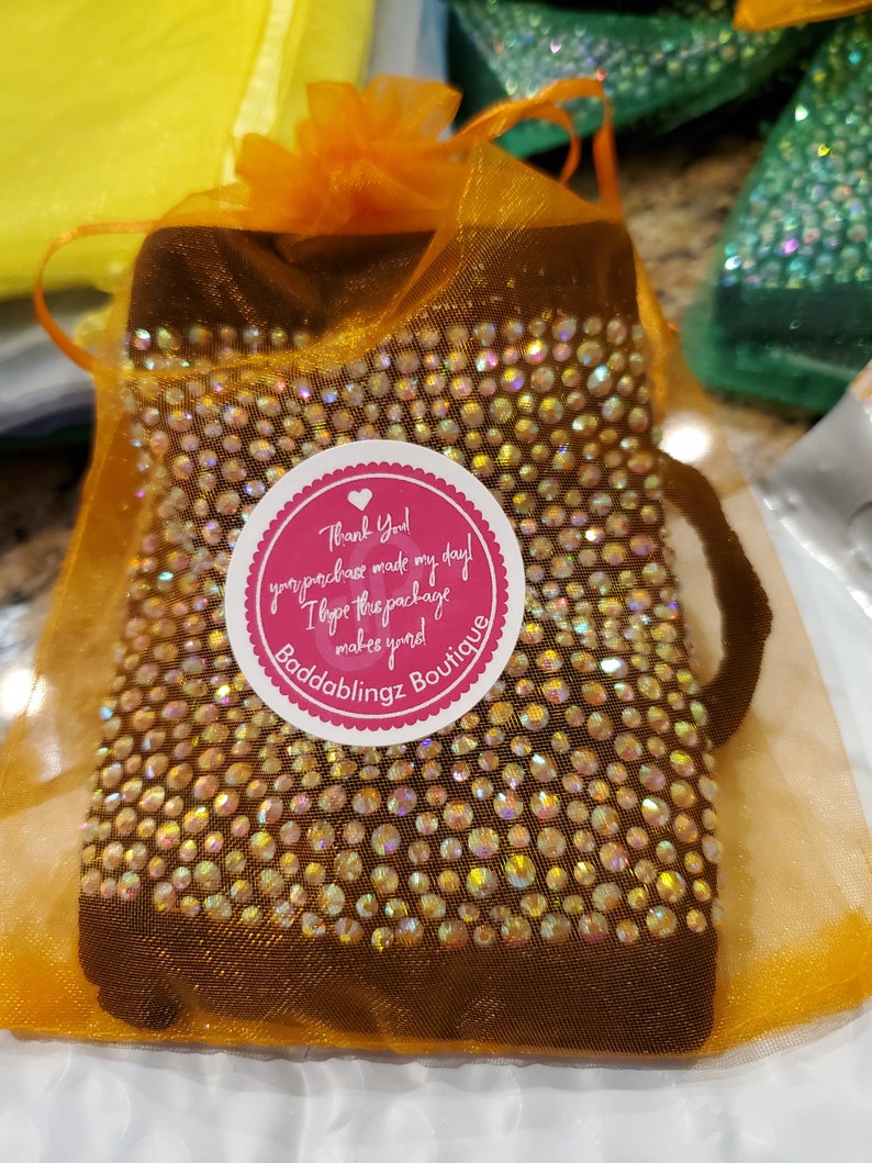 May include: A brown pouch with a sparkly design is in a bright orange organza bag. The pouch has a round sticker with the text "Thank You! Your purchase made my day! I hope the package makes you! Baddablingz Boutique" in pink and white text.