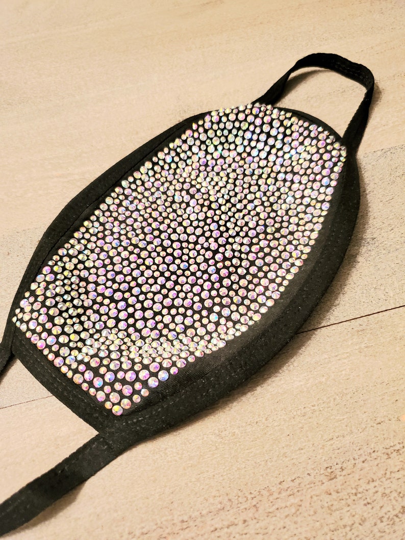 May include: A black face mask covered in iridescent rhinestones. The mask has elastic ear loops.