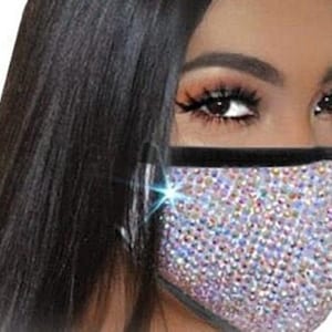 May include: A face mask covered in iridescent rhinestones with a black elastic band. The mask has a black trim around the edges.