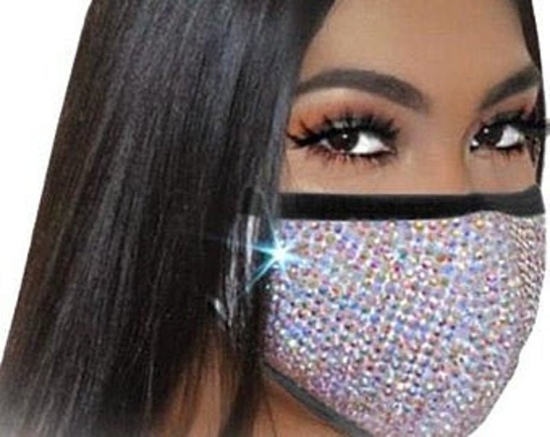 May include: A face mask covered in iridescent rhinestones with a black elastic band. The mask has a black trim around the edges.