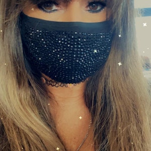 May include: A woman with long brown hair wearing a black face mask with rhinestones. The mask covers her nose and mouth.
