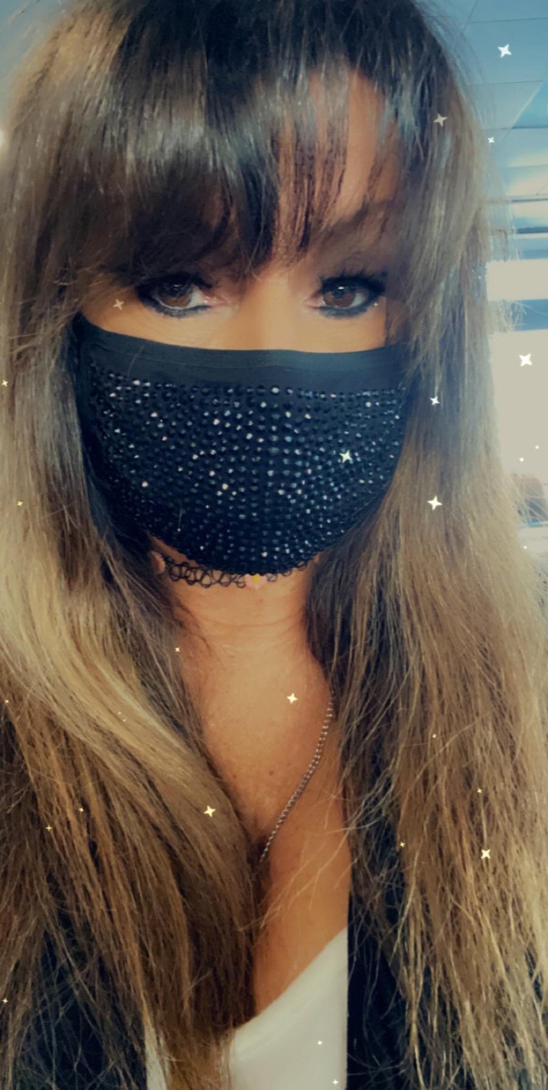 May include: A woman with long brown hair wearing a black face mask with rhinestones. The mask covers her nose and mouth.