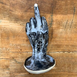 Könnte beinhalten: Schwarze Keramik-Handskulptur mit einem weiß bemalten Mittelfinger und einem weiß bemalten Symbol auf der Handfläche.