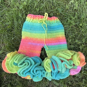 Op de afbeelding: Een kleurrijke, handgehaakte broek. De broek heeft een regenboog aan kleuren, waaronder roze, oranje, geel, groen en blauw. De onderkant van elke broekspijp heeft een gefranjerde rand. Een groen trekkoord zit in de taille.