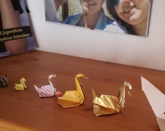 Może przedstawiać: Pięć origami w kształcie łabędzi wykonanych z papieru, jeden jest biały z ręcznie napisanym Sudoku, pozostałe są złote i różowe. Łabędzie znajdują się na drewnianej powierzchni.