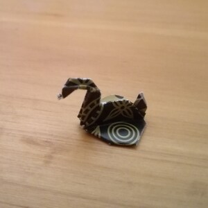 May include: A small origami swan made from paper with a black and gold floral pattern.
