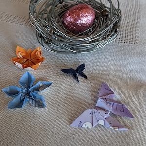 Puede incluir: Un nido de alambre con un huevo de chocolate envuelto en papel de aluminio rosa en el centro. Cuatro mariposas de origami están dispersas alrededor del nido, incluyendo una mariposa azul, una mariposa morada y dos mariposas naranjas.