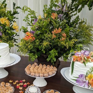 May include: A table setting with two white cakes decorated with flowers, surrounded by small brown and white candies, and a clear glass candle holder. The table is decorated with origami paper butterflies in shades of orange, yellow, and red.