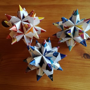May include: Three origami stars made from colorful paper. The stars are white with different colored patterns on each point. The stars are sitting on a wooden surface.