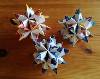 Puede incluir: Tres estrellas de origami hechas de papel de colores. Las estrellas son blancas con diferentes patrones de colores en cada punta. Las estrellas están sobre una superficie de madera.