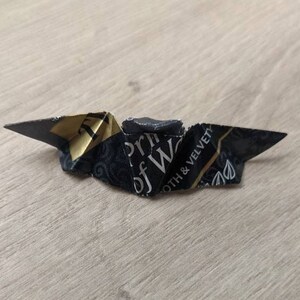 Puede incluir: Una figura de origami plegada con papel estampado en negro y dorado. El papel tiene el texto "Prince of Wales" y "Smooth & Velvety". El origami est&aacute; sobre una superficie de vetas de madera clara.