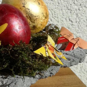 May include: A gray stone bowl filled with moss and decorated with two Easter eggs, one red and one gold, and two origami butterflies made from candy wrappers. The butterflies are orange and yellow.
