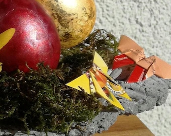 Może przedstawiać: Szara kamienna miska wypełniona mchem i ozdobiona dwoma jajkami wielkanocnymi, jednym czerwonym i jednym złotym, oraz dwoma origami motylami wykonanymi z papieru po cukierkach. Motyle są pomarańczowe i żółte.