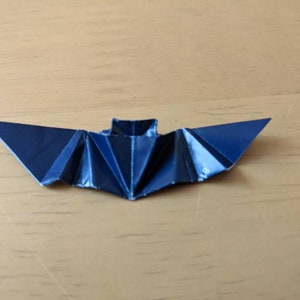 Puede incluir: Un murci&eacute;lago de origami azul oscuro, hecho de papel plegado, descansa sobre una superficie de madera marr&oacute;n claro. Las alas del murci&eacute;lago est&aacute;n extendidas, mostrando los intrincados pliegues y &aacute;ngulos agudos del papel. El cuerpo del murci&eacute;lago es una forma rectangular.
