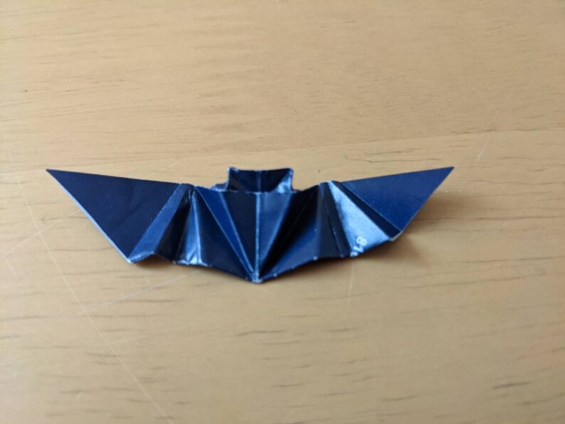 Puede incluir: Un murci&eacute;lago de origami azul oscuro, hecho de papel plegado, descansa sobre una superficie de madera marr&oacute;n claro. Las alas del murci&eacute;lago est&aacute;n extendidas, mostrando los intrincados pliegues y &aacute;ngulos agudos del papel. El cuerpo del murci&eacute;lago es una forma rectangular.