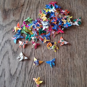 May include: A collection of colorful origami butterflies made from recycled paper. The butterflies are arranged in a pile on a wooden surface.