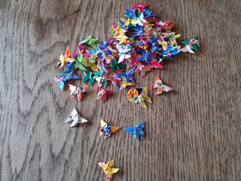 May include: A collection of colorful origami butterflies made from recycled paper. The butterflies are arranged in a pile on a wooden surface.