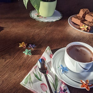 May include: A wooden table with a cup of hot chocolate, a plate of chocolate squares, a green plant in a pot, and a napkin with origami flowers.