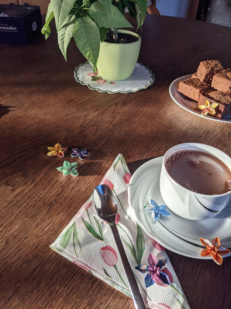 May include: A wooden table with a cup of hot chocolate, a plate of chocolate squares, a green plant in a pot, and a napkin with origami flowers.
