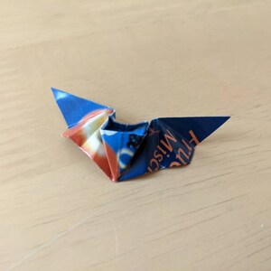 Puede incluir: Un p&aacute;jaro de origami plegado con papel estampado azul y naranja. El papel tiene texto. El p&aacute;jaro de origami est&aacute; sobre una superficie de madera marr&oacute;n claro.