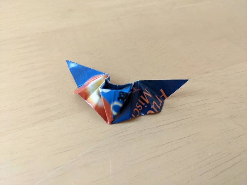 Puede incluir: Un p&aacute;jaro de origami plegado con papel estampado azul y naranja. El papel tiene texto. El p&aacute;jaro de origami est&aacute; sobre una superficie de madera marr&oacute;n claro.