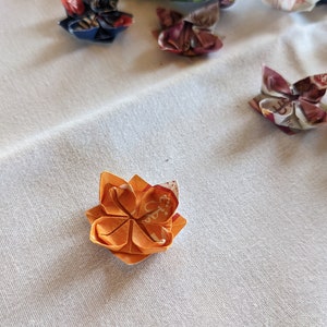 May include: A small orange origami flower with white and red accents. The flower is folded from paper and has a five-petal design.