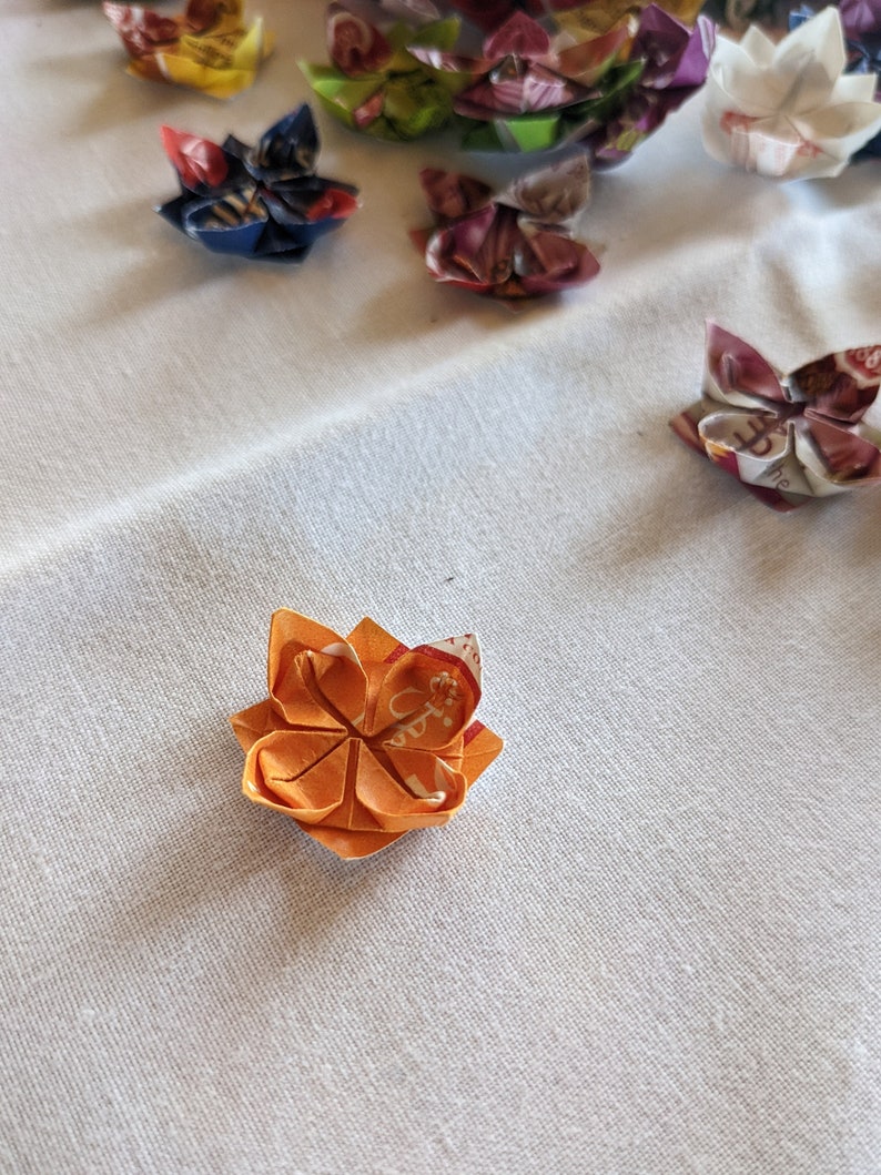 May include: A small orange origami flower with white and red accents. The flower is folded from paper and has a five-petal design.