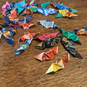 Puede incluir: Una colecci&oacute;n de murci&eacute;lagos de origami hechos de papel reciclado colorido. Los murci&eacute;lagos son de varios colores, incluyendo rojo, azul, verde y negro, y est&aacute;n dispuestos sobre una superficie de madera. Las alas est&aacute;n extendidas.