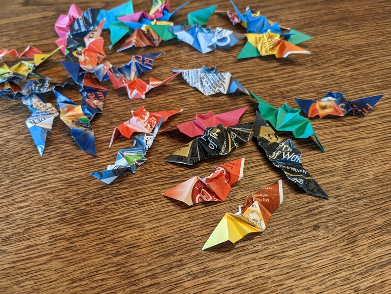 Puede incluir: Una colecci&oacute;n de murci&eacute;lagos de origami hechos de papel reciclado colorido. Los murci&eacute;lagos son de varios colores, incluyendo rojo, azul, verde y negro, y est&aacute;n dispuestos sobre una superficie de madera. Las alas est&aacute;n extendidas.