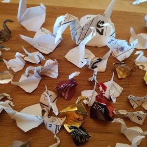 May include: A collection of origami paper cranes made from various printed papers, including a calendar page, a candy wrapper, and a magazine page. The cranes are arranged on a wooden surface.