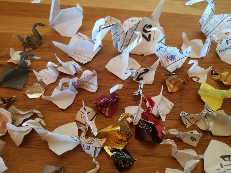 May include: A collection of origami paper cranes made from various printed papers, including a calendar page, a candy wrapper, and a magazine page. The cranes are arranged on a wooden surface.
